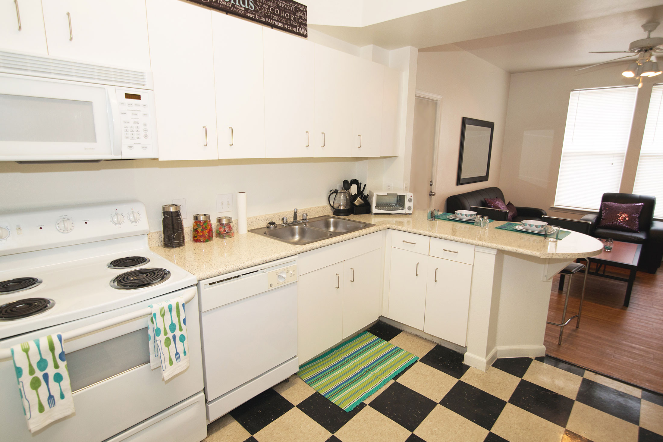 Kitchen with checkerboard floor and white cabinets and appliances including a range, dishwasher and microwave with stainless steel double basin sink, open to the Living Area.