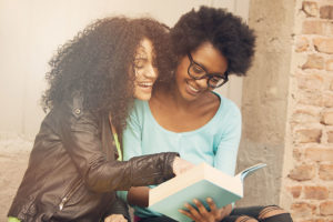 Two black friends looking and laughing at a book outdoors. 
